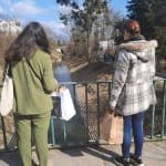 Dwie kobiety z torbami zakupowymi stoją na kładce i patrzą się na nurt rzeki. Są odwrócone tyłem do obiektywu.