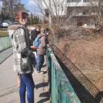 Dwie panie i mężczyzna stoją na kładce zwróceni bokiem do aparatu. Patrzą przed siebie. W tle biały dwupiętrowy budynek i szary samochód.