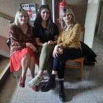 Trzy kobiety - jedna w wieku średnim i dwie młode siedzą na krzesłach na korytarzu i pozują do zdjęcia. Za nimi wisząca na ścianie gaśnica i tablica z napisem: Zakaz palenia na terenie obiektu.