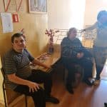 Dwoje ludzi - mężczyzna i kobieta siedzą na krzesłach obok stolika z ułożonymi czasopismami i flakonem kwiatów. Obok siedzącej kobiety stoi druga kobieta. Na ścianie wisi duży obraz i dwie instrukcje przeciwpożarowe.