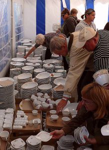 Ludzie pochylaja się do skupiska talrzy i filiżanek. Są w namiocie.