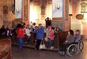 Ludzie są w kościele. Niektórzy siedzą w ławach. Są dwie kobiety na wózkach inwalidzkich.