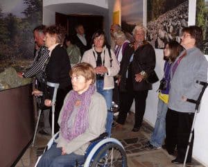 Ludzie stoją w pomieszczeniu. Niekótrzy są o kulach lub wózkach. Ich twarze są sierowane na wporst obrazów które wiszą na scianie