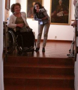 Kobiety stoją przed schodami. Jedna jest na wózku dryga pochyla sie w jej kierunku i wyciaga rękę przed siebie