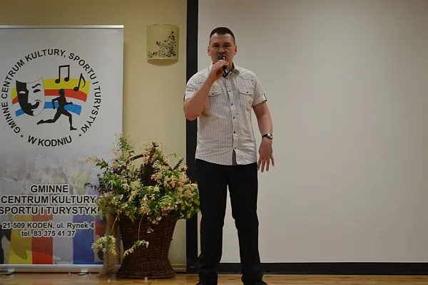 Mężczyzna stoi na środku sali. Trzyma mikrofon w dłoni. PO jego lewej stronie w tle jest baner reklamowy stojący.