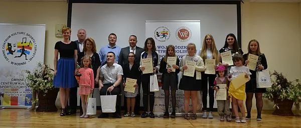 Ludzie stoją na środku sali w grupie. Pozują do zdjęcia. W dłoniach trzymają dyplomy i torby upominkowe. Uśmiechaja sie do zdjecia.