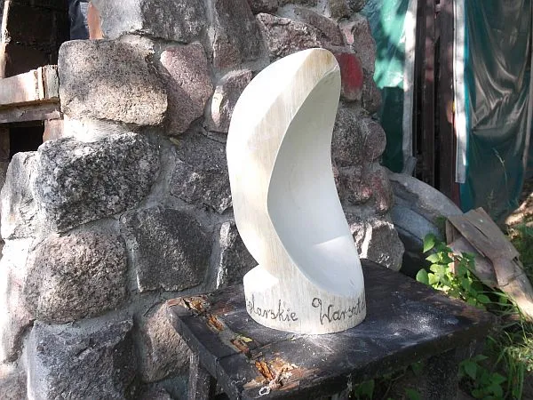 Rzeźba o obłym kształcie. Jest na tle murku kamiennego. Stoi na taborecie.