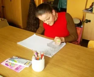 Kobieta siedzi przy stole. W dłoni trzyma kredkę. Dłoń jest na kartce. Obok kartki są kolorowe kredki