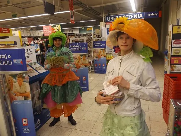 Dwie kobiety w strojach stoją w sklepie. Kobieta po prawej stronie ma na sobie kapelusz. KObieta po lewej stronie ma na sobie zielone włosy i kolorową spódnicę.
