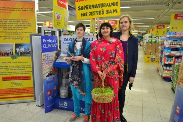 Trzy kobiety pozują do zdjęcia. Kobieta pośrodku trzyma koszyk i jest ubrana w sukienkę w kwiaty. Obok stoi kobieta w niebieskim szalu. Są one w sklepie spożywczym.
