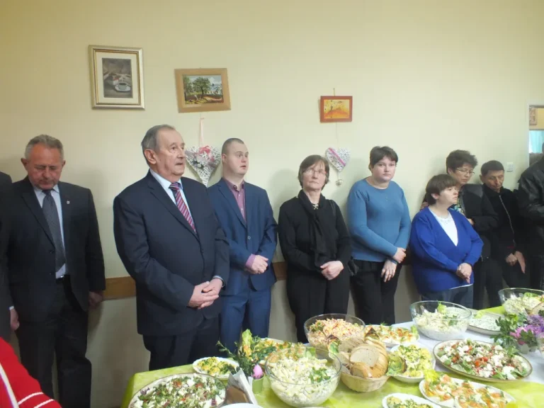 Ludzie wizytowo ubrani stoja. Przed nimi stół zastawiony jedzeniem.