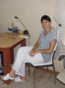 Kobieta siedzi na krześle. Obok stół na którym są drobiazgi. Kobieta uśmiecha się do zdjęcia.