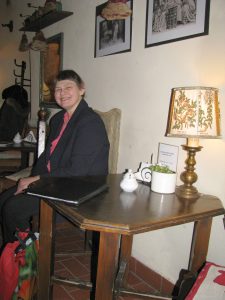 Kobieta siedzy na krzesle. Rękę opiera o stolik. Na stoliku jest czarna teczka. Kobieta uśmiecha sie do zdjęcia.