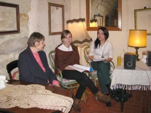 Ludzie siedzą na krzesłach. Twarze mają zwrócone ku sobie. Uśmiechaja sie do siebie. W tle puste ramy obrazów wiszące na ścianach.