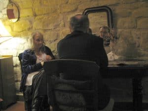 Meżczyzna siedzi przy biurko. Nad biurkiem wisi lustro które odbija jego twarz. Po lewej stronie jest kobieta, która ma twarz zwróconą w strone mezczyzny.