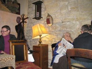 Ludzie siedzą przy stołach w kawiarni. Ich twarze sa zwrócone ku sobie. Jedna z kobiet trzyma gazete w dłoniach.