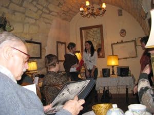 Ludzie są w kawiarni. Niektórzy siedzą. W tle dwie kobiety, które stoją. Ich twarze sa zwrócone ku sobie. Jeden z mężczyzn siedzących przy stoliku trzyma teczkę w dłoniach.