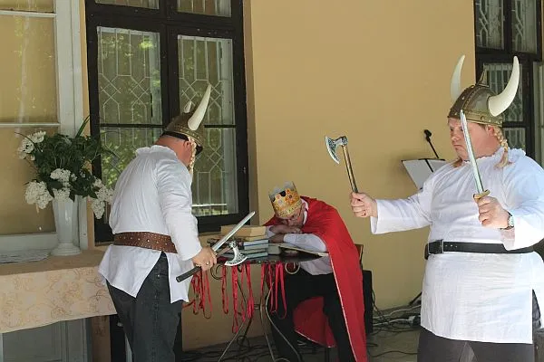 dwóch mężczyzn jest przebranych za wikingów. Na głowach mają hełmy z białymi rogami po bokach. W dłoniach trzymają siekiery i miecze. W tle mężczyzna przebrany za króla.