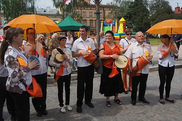 Ludzie stoją w grupie na deptaku w mieście. Niektórzy z nich trzymają w dłoniach pomarańczowe parasole a inni bębenki owinięte pomarańczowymi chustami.