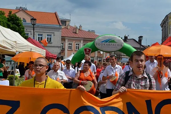 Ludzie idą deptakiem w korowodzie. Dwóch mężczyzn trzyma w dłoniach materiałowy pomarańczowy długi baner. W tle niektórzy trzymają pomarańczowe parasolki albo bębenki.