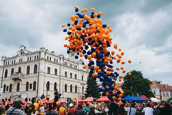 ludzie stojący na deptaku w mieście. Balony wypełnione helem w dwóch kolorach - pomarańczowym i granatowym zostały wypuszczone do nieba. Wznoszą się.
