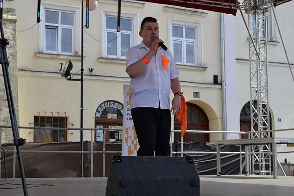 Mężczyzna stoi na scenie rozłożonej na deptaku miasta. Do białej koszuli ma pomarańczowe dodatki dodane.