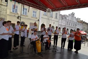 Ludzie stoja w grupie na scenie . Mężczyzna w środku trzyma bęben afrykański. Pozostałe osoby klaszczą w ręce albo trzymają grzechotki i tamburyny w nich.