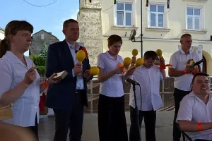 Ludzie stoją w grupie. W dłoniach trzymają grzechotki. Przed nimi jest mikrofon stojący.