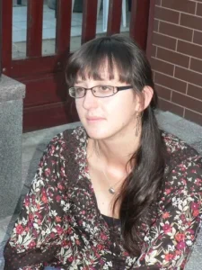 Na zdjeciu jest kobieta, uśmiecha sie do zdjecia. Jej twarz skierowana jest pzed siebie.