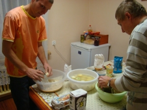 Ludzie stoja nad stołem. Pochylają sie nad nim. Na stole są miski z jedzeniem.