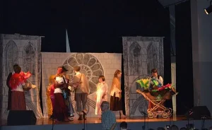 Na scenie jest scenografia kościoła. Przed nim stoi grupa osób. Po prawej stronie jest wózek wypełniony kwiatami, a z nim kobieta.