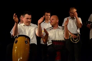 Dwóch mężczyzn wykonuje gest klaskania. Obaj mają przed sobą bębny afrykańskie. Za nimi stoją inni mężczyźni.
