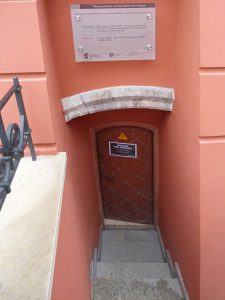 Jest wejście do sutereny. Są schody do wejścia. Budynek jest koloru brzoskiwniowego.