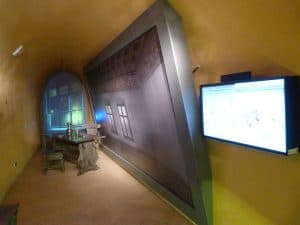 Sala z dyżym ekranem multimedialnym przedstawiajacym wnętrze pokoju. Przy ekranie jest stól z krzesłem. W tle okno.