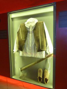 Za podświetlonym ledami szkle w ścianie jest strój ziemianina, biała koszula, brązowa narzuta, trzewiki i tasak.