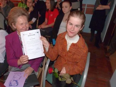 Kobiety siedza. W dłoniach razem trzymaja dyplom. Uśmiechają sie do zdjęcia.