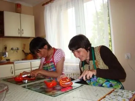 Kobiety pochylają sie nad stołem. Kroją czerwoną paprykę. Uśmiechaja sie przy tym. W tle szafki kuchenne,