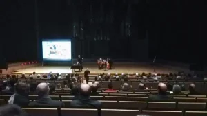 Ludzie siedzą na widowni. Na scenie są ludzie z instrumentami.