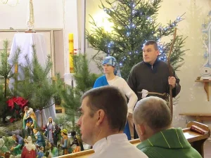 Mężczyzna trzyma w dłoni laskę. Obok niego osoba z nakrytą w torban głowa. W tle za nimi choinki. Przed mężczynami ludzie siedzą w ławach.