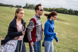 Ludzie idą deptakiem. Dwoje z nich w dłoniach ma laski dla niewidzących. W tle trwanik. Jest pochmurno.