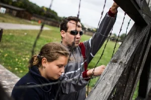 Ludzie dotykają druciane kolczaste ogrodzenie. Ich twarze są zwrócone na wprost. W tle jest trawnik i drzewa,