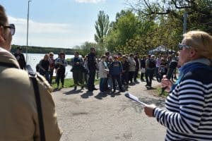 Grupa osób stoi wśród drzew. Po prawej stronie stoi kobieta z mikrofonem, a w ręce trzyma teczkę i jest bokiem do obiektywu. Po lewej stronie stoi osoba, która jest ucięta przez obiektyw.