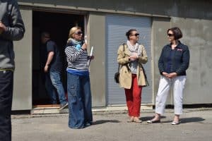 Przed budynkiem stoją trzy kobiety. Kobieta po lewej stronie trzyma mikrofon blisko twarzy. Za nią jest mężczyzna stojący w środku pomieszczenia, tyłem do obiektywu.