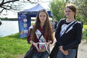 Dwie dziewczyny stoją przed obiektywem. Po lewej stronie dziewczyna patrzy wprost w niego i trzyma puszkę. Dziewczyna po prawej stronie ma głowę skierowaną w lewą stronę. Za nimi jest namiot i baner z napisem: “Szkoła Policealna”.