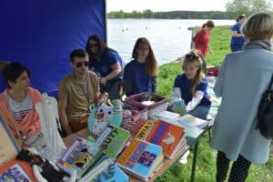 Ludzie siedzą pod namiotem na krzesłach. Na stołach przed nimi leżą różne książki zrobione własnoręcznie. Po prawej stronie stoi tyłem kobieta.