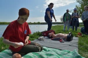 KObieta po lewej stronie klęczy na kocu i trzyma w ręku gazik. Obok niej leży fantom dziecka ucięty przez obiektyw, a po prawej stronie fantom dorosłego człowieka. Po prawej stronie stoi czwórka osób.