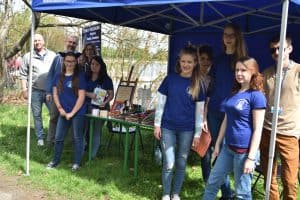 Grupa osób pozuje do zdjęcia. Mają na sobie niebieskie koszulki. Na stołach leżą różne wyroby ręczne.