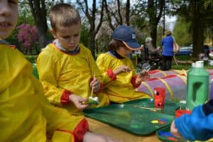Przy stołach siedzą małe dzieci. Na stole leżą tacki na których jest różnokolorowa farba. Chłopiec po lewej stronie trzyma pędzel i uformowaną masę solną. Po prawej stronie w oddali jest starsza pani na wózku inwalidzkim a obok stoi inna kobieta. Są one tyłem do obiektywu.