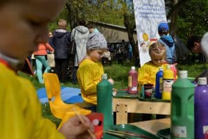 Przy stoliczkach siedzą dzieci, które mają tacki z farbą. Na stolikach stoją butelki z różnymi kolorami farb. Po prawej stronie baner z napisem: “Niepubliczne przedszkole dla dzieci niepełnosprawnych, Krasnystaw, ul. Kwiatowa 2”.