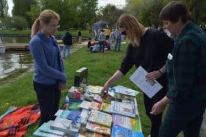 Na stole leżą różne książki. Po lewej stronie stoi kobieta, a po prawej dwie kobiety nachylają się nad stołem. Za nimi stoi grupa osób.